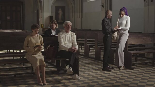 Wide shot of three Caucasian parishioners sitting on pews in Lutheran church while African-American pastor talking to young African woman nearby