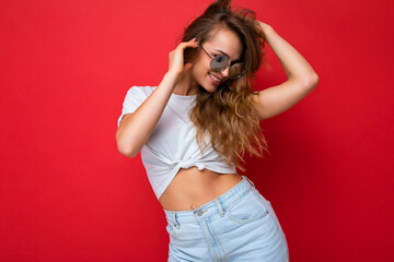 Fototapeta premium Photo shot of beautiful happy young blonde woman wearing casual clothes and stylish sunglasses isolated over colorful background and having fun