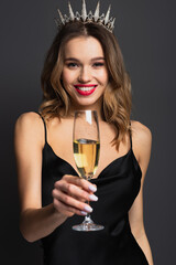joyful young woman in black slip dress and tiara holding blurred glass of champagne on grey © LIGHTFIELD STUDIOS
