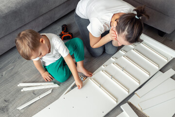 Cute adorable caucasian toddler boy kid sit on floor and help mom assembling furniture shelf with...