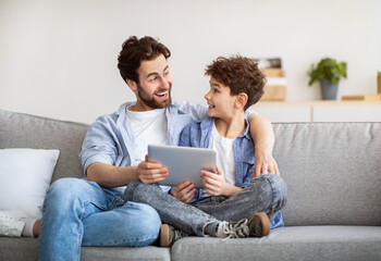 Family weekend. Excoted dad and son using tablet computer, surfing internet while spending time...
