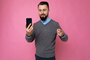 Handsome happy cool young brunette unshaven man with beardwearing stylish grey sweater and blue shirt standing isolated over pink background wall holding smartphone and using phone looking at gadjet