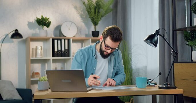 Handsome in good mood confident smart 30-aged bearded man sitting at the table in home office and writing down important notes into paper report