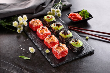 International maki sushi roll on black slate plate. Served with wooden chopstick, green wasabi and pickled ginger. Japanese set with maki sushi roll on dark stone background..
