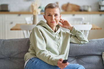 Movie time. Portrait of beautiful middle aged woman with remote control in hand watching tv while sitting and relaxing on sofa, spending leisure time at home. Selective focus on senior female