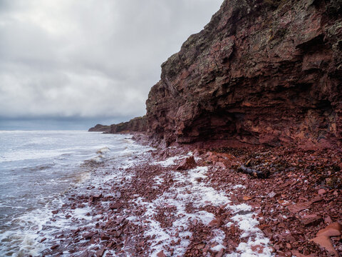 A cliff above the sea with a narrow coastline. Waves with white foam roll on the rocky shore. Tersky coast