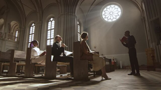 Panning of African-American pastor wearing black trousers, shirt and white collar standing in Lutheran church and preaching sermon to several diverse parishioners