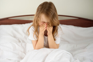 A little girl sits and sneezes on the bed among the blankets and pillows. House dust mite allergy