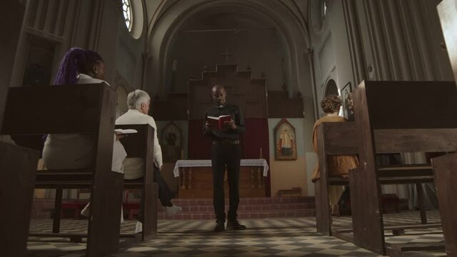 Zoom out of African-American clergyman wearing black clothes and white collar standing in Lutheran church with Holy Bible in hands and speaking in front of several parishioners