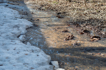 A spring stream of melt water flows over the muddy ground with the remnants of snow.