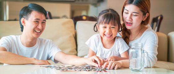 Obraz premium Banner photo of Asian family teach their daughter to saving money and putting coins into glass bank spend a quality time together. Asian family, relationship, happiness and investment concept