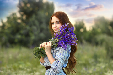 Banner with a stunning young girl looking at the camera with a smile, a redhead holding a bouquet of lilac lupins in her hands. Background blue sky and sunset. Nature. Summer concept. Copy space