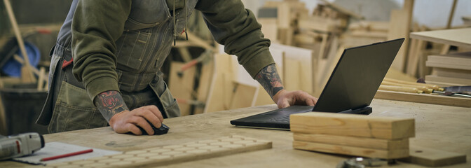 Concentrated adult craftsman checking information on laptop while working in carpentry workshop....