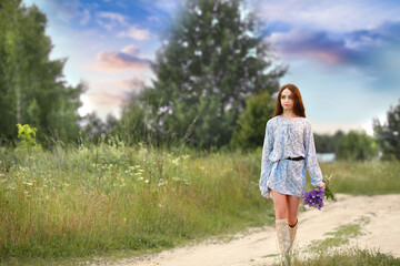 A beautiful girl in a blue dress and summer boots walking along the road where there are wildflowers and a meadow with a bouquet of lilac lupins. Sunset background, summer landscape. Copy space