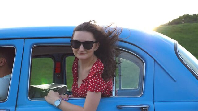 Happy girl in sunglasses leaning out of retro car window and enjoying trip. Young woman looking out window of moving old auto on sunny day. Travel and freedom concept. Slow motion Close up