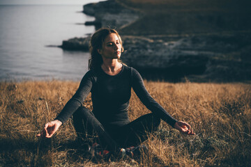 Young woman in black shirt practices yoga asanas on high rocky coast above sea at bright sunlight and soft wind in tranquil morning