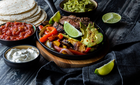 Close up view of a skillet filled with ingredients used in making fajitas and surrounded by toppings and tortillas.