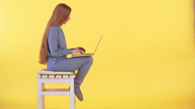 A woman with long ginger hair is sitting in casual costume and socks on the chair and working online on the laptop. Freelancer and businesswoman. Yellow background
