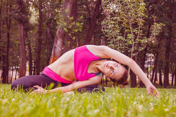 Naklejka na ściany i meble European girl in a light tracksuit doing yoga in the park in the afternoon in the summer. Gymnastics in nature. Thin woman outdoor