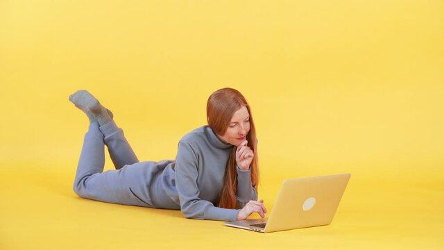 Young woman with long ginger hair is lying on the floor and browsing the Internet on the laptop. Yellow background