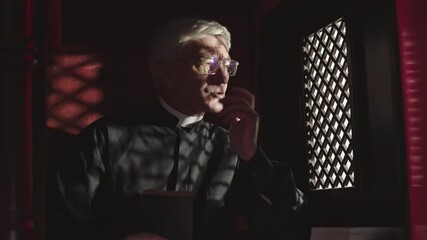 Medium shot of senior Caucasian priest wearing black robe, white collar and eyeglasses sitting in confessional near window, listening to confession and the reading Holy Bible
