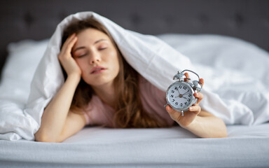 Sleepy young lady lying under blanket in bed with alarm clock, having problem waking up in morning,...