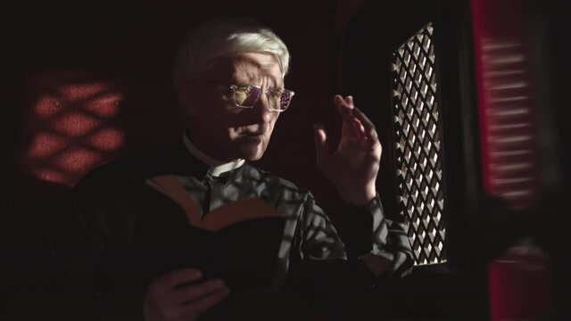 Medium shot of old Caucasian priest wearing black robe, white collar and eyeglasses sitting in confessional with Holy Bible in hands and listening to confession