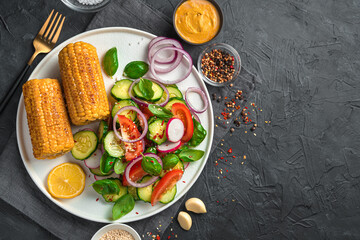 Vegetable salad with tomatoes, cucumbers and radishes and heads of corn in a white flat plate on a black background with space to copy.