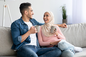 Cheerful arab couple posing on sofa, holding mugs with drinks