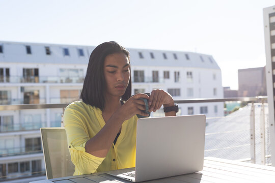 Mixed race transgender woman sitting at table on sunny roof terrace using laptop drinking coffee