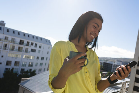 Smiling mixed race transgender woman standing on sunny roof terrace holding coffee using smartphone