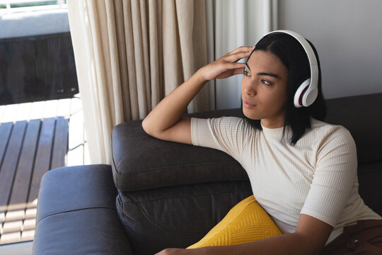 Happy mixed race transgender woman relaxing in living room sitting on couch with headphones