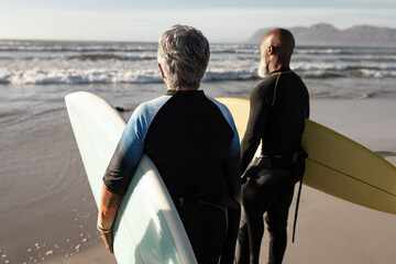 Diverse senior couple on beach holding surfboards looking out to sea