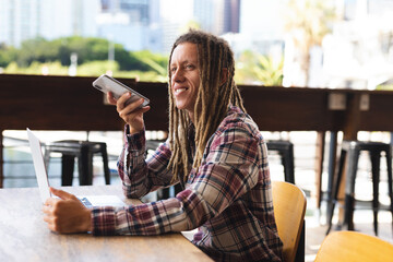 Smiling mixed race man with dreadlocks sitting at table outside cafe talking on smartphone