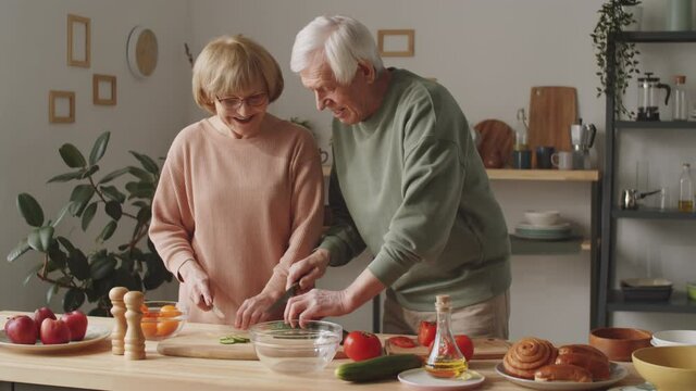 Senior wife and husband cutting fresh vegetables and having discussion while cooking dinner together in kitchen