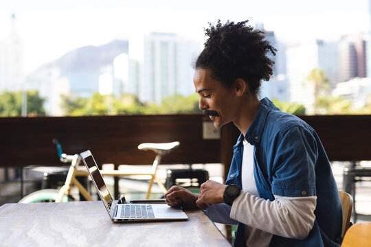 Smiling mixed race man with moustache sitting at table outside cafe using laptop