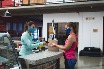 Female receptionist and customer wearing masks disinfecting hands over the counter at gym