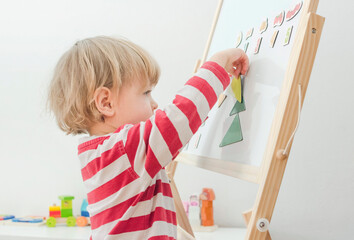 Cute little child playing at a magnetic board, learning letters, numbers and geometric shapes at home. Educative and funny indoor activity for early development.