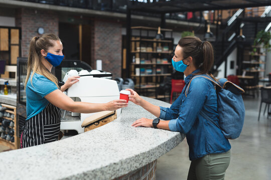Two happy caucasian women wearing masks passing cup of coffee over counter