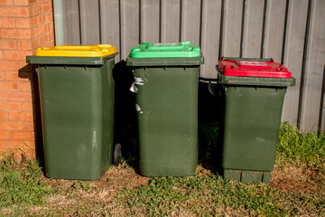 Australian garbage wheelie bins with red and yellow lids for general and recycling household waste and green lid for garden waste near the resedential building fence