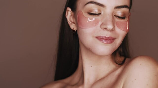 Facial care. Skin treatment. Anti aging procedure. Portrait of playful woman with nude makeup bare shoulders applying pink hydrogel eye patches winking isolated on brown background.