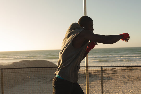 African american man exercising outdoors shadowboxing with wrapped hands on beach at sunset - Powered by Adobe
