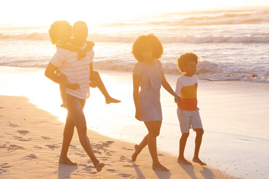 African american parents and two children walking holding hands and piggy backing at the beach