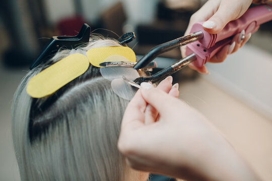 Hairdresser female making hair extensions to young woman with blonde hair in beauty salon. Professional hair extension