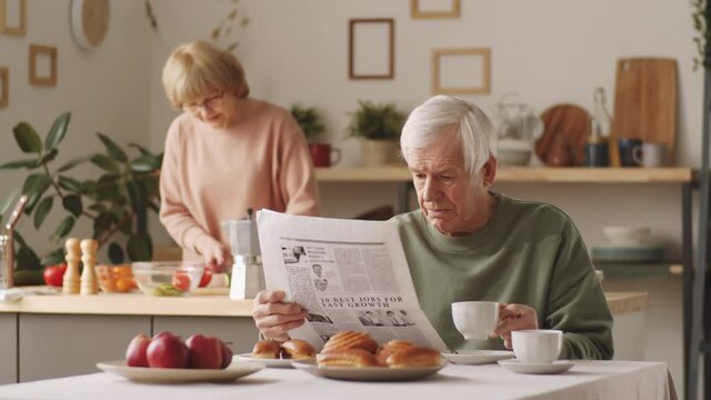 Elderly Caucasian man drinking coffee and reading newspaper at kitchen table while his wife cooking meal in background
