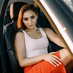 A beautiful young girl sits in a car in the evening in the sunset summer sun in an empty parking lot