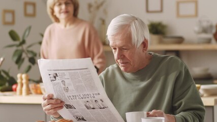 Tilt up shot of senior man reading newspaper at kitchen table, drinking coffee and speaking with wife while she cooking in background - Powered by Adobe