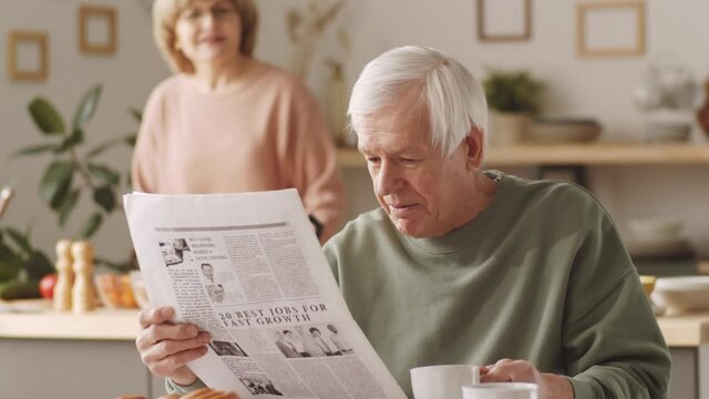 Tilt up shot of senior man reading newspaper at kitchen table, drinking coffee and speaking with wife while she cooking in background