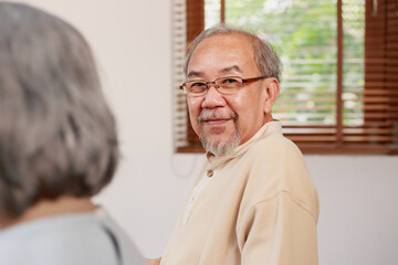 Obraz premium Happy Asian senior man sitting by white hair elderly woman looking at camera with smiley face by the window at home. Friendly Asian retired people with beard wearing glasses with happy smile indoor