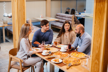 A group of young cheerful friends is sitting in a cafe talking and eating pizza. Lunch at the pizzeria.